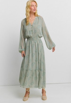 Femme portant une longue robe vert clair à motifs, manches longues, corsage boutonné, taille smockée, et chaussures compensées beiges à lacets à la cheville, debout sur un sol blanc.