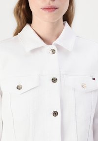 Close-up af en person iført en hvid denimjakke med metal knapper og et lille rødt, hvidt og blåt Tommy Hilfiger-logo på lommen.