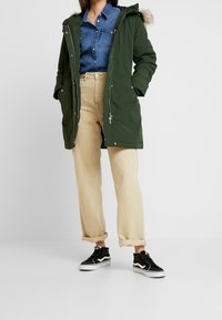 Grüne Parka mit fellgefütterter Kapuze, Reißverschluss und Druckknöpfen, getragen über einem Jeanshemd, kombiniert mit beigen Hosen und schwarzen Sneakers.