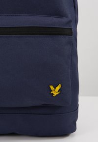 Sac à dos bleu marine en tissu texturé, avec un logo d'aigle jaune et une poche avant zippée. Forme rectangulaire, conception durable.