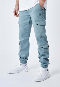 Lichtblauwe cargobroek met elastische boorden, meerdere zakken en een klein geborduurd logo. Draagt met witte sneakers.