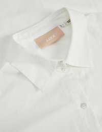 Camisa de color blanco con cuello, de textura suave, mangas largas y una etiqueta etiquetada con "JJXX" en rosa suave con texto blanco. Dos botones blancos visibles.