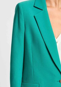 Blazer turquesa con un corte ajustado, solapas afiladas, hombros estructurados y un detalle de un solo botón. Textura de tela suave.
