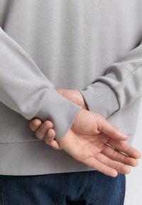 Lichtgrijze sweater met ronde hals en geribbelde manchetten, gemaakt van een textuurstof en met een relaxed fit, gedragen met donkerblauwe jeans. De handen zijn op de rug samengevoegd.