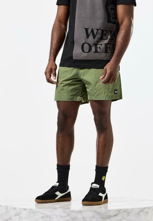 Grønne nylon shorts med en tekstureret overflade, parret med en sort grafisk T-shirt med hvid tekst. Sorte sneakers med hvide detaljer.