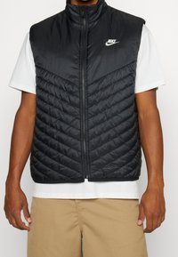 Svart pufferväst med hög krage, dragkedjeslutning och vingmönstrad quiltad design. Logotyp på vänster bröst, bärs över en vit skjorta.