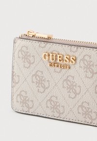 Bézs színű, texturált pénztárca párnázott mintával, aranyozott, dombornyomott „GUESS EST 1981” logóval és fémből készült cipzáras záródással.