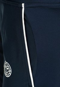 Close-up van marineblauwe broek met een zijzak, witte verticale streep en een wit cirkelvormig abstract logo nabij de zak.