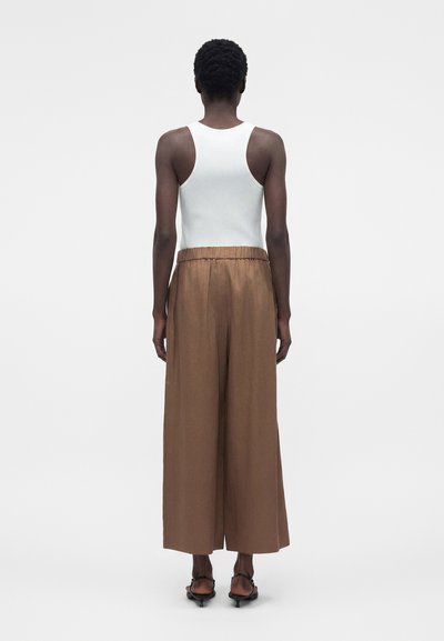 Άτομο που στέκεται πλάτη προς το φακό, φοράει λευκό αμάνικο ριμπ πλεκτό τοπ, φαρδιά καφέ παντελόνια και μαύρα σανδάλια με λουράκια και χαμηλό τακούνι.