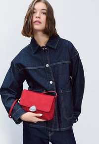 Džínová bunda tmavomodrej farby so striebornými gombíkmi a prešitím. Drží malú červenú semišovú kabelku so striebornou sponou.