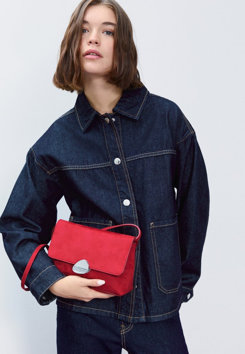 Džínová bunda tmavomodrej farby so striebornými gombíkmi a prešitím. Drží malú červenú semišovú kabelku so striebornou sponou.