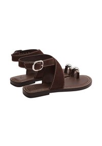 Brązowe zamszowe sandały na płaskiej podeszwie, z krzyżującymi się paskami, zapięciem na kostce oraz dwiema srebrnymi dekoracyjnymi kulkami z przodu. Widoczne przeszycia.