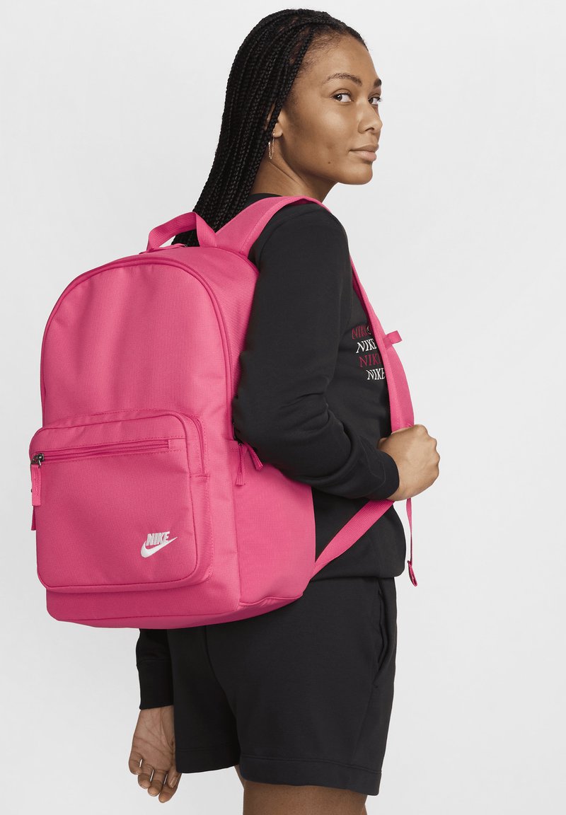 Ροζ σακίδιο Nike με υφή, μπροστινή τσέπη με φερμουάρ, ρυθμιζόμενους ιμάντες και λεπτομέρεια λογότυπου. Μοντέλο που φοράει μαύρη φούτερ.