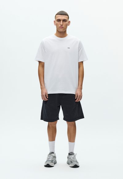 Λευκό βαμβακερό T-shirt με μικρό μαύρο λογότυπο, συνδυασμένο με μαύρο σορτς. Γκρι αθλητικά παπούτσια με λευκές κάλτσες. Μινιμαλιστικός σχεδιασμός και ουδέτερα χρώματα.