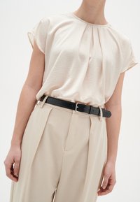 Lys beige bluse med foldet halsudskæring, korte ærmer, kombineret med beige højtaljede bukser og et sort læderbælte.