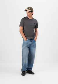 Graues T-Shirt mit kurzen Ärmeln und kleinem Logo, kombiniert mit locker sitzenden, hellblauen Jeans und schwarzen Sneakern. Olivgrüne Mütze.