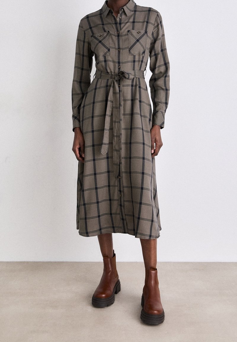 Femme portant une robe midi à carreaux beige et noire avec une ceinture nouée et des bottines marron, debout sur un sol clair contre un mur blanc.