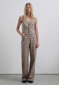 Monos de cuadros marrones con parte superior de chaleco ajustado, seis botones negros, pantalones de pierna ancha y bolsillos laterales, fabricado en tejido ligero.