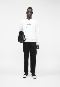 Jovem vestido com um hoodie branco com o logotipo "Guess Jeans", calças pretas, ténis pretos, segurando uma mala preta contra um fundo simples.