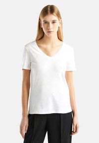 Giovane donna con capelli biondi lisci, che indossa una semplice maglietta bianca a V con maniche corte e pantaloni neri, in piedi contro uno sfondo bianco.
