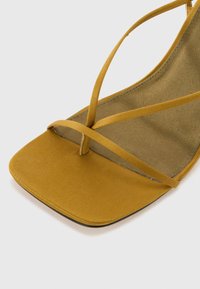 Sandal jaune avec une texture douce et mate, présentant de fines lanières croisées et un design à bout carré, reposant sur un coussinet beige.