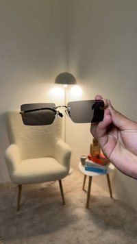 Graue Sonnenbrille mit quadratischen Gläsern und silbernen Metallakzenten, von einer Hand vor einem hellen Hintergrund mit weichen Texturen gehalten.