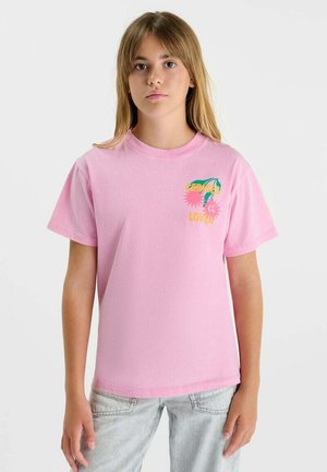 Fille aux cheveux blonds raides portant un t-shirt rose avec un texte floral coloré sur la poitrine et un jean gris clair, debout devant un fond blanc.