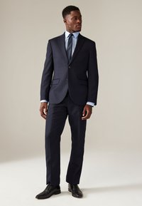 Tumesinine kohandatud ülikond koos istuva jopeta, kahe nupu, sälgfööside, sobivate pükste ja all helerohelise särgiga. Mustad nahast kingad.