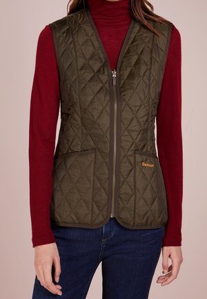 Femme portant une veste matelassée zippée vert olive sur un col roulé bordeaux manches longues et un jean bleu foncé.