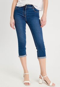 Pantalons capri en denim, bleu foncé, avec des ourlets roulés et un design à cinq poches. Associés à des sandales à talons blocs blanches. Tissu lisse et texturé.