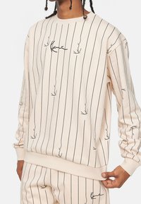 Felpa beige con righe verticali nere e disegni minimalisti. Presenta un collo a giro e polsini e orlo a coste.