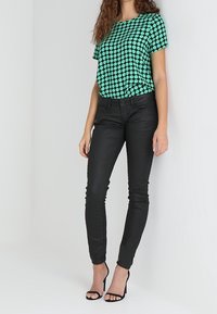Mujer vestida con una blusa corta de manga en color verde azulado con puntos, pantalones negros ajustados y sandalias de tacón negro, de pie frente a un fondo claro.