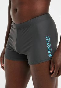 Dunkelgraue Badeshorts aus glattem, elastischem Material. Mit einem blauen "PROTEST"-Logo an der Seite und einem taillierten Schnitt.
