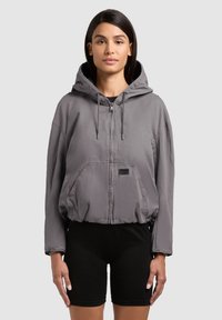 Femme portant une veste à capuche grise zippée et un short de cyclisme noir, debout devant un fond clair uni.