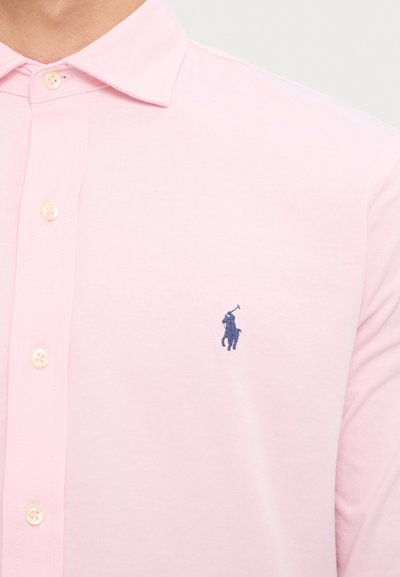 Světle růžová polo tričko vyrobené z bavlny, s tmavě modrým vyšitým logem na hrudi a knoflíky, které ladí s barvou látky.