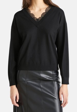 Pull noir à manches longues et décolleté en V, orné d'une délicate dentelle noire au niveau du col, porté avec une jupe en cuir noire.