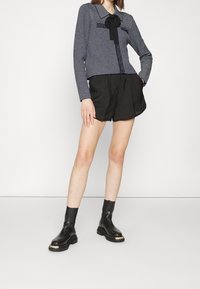 Tummansininen ja valkoinen kirjailtu paita, jossa on solmuke, yhdistetty mustiin shortseihin ja mustiin nilkkasandaaleihin, joissa on paksu pohja ja kultaiset yksityiskohdat.