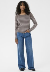 Maglione a righe grigie con dettagli metallici, abbinato a jeans in denim blu a gamba larga. Il modello sta in piedi, con le mani nelle tasche, su uno sfondo semplice.