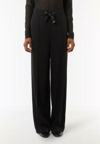 Czarne spodnie o szerokich nogawkach z wiązanym pasem, wykonane z lekkiego materiału. Posiadają plisowania i gładką teksturę.