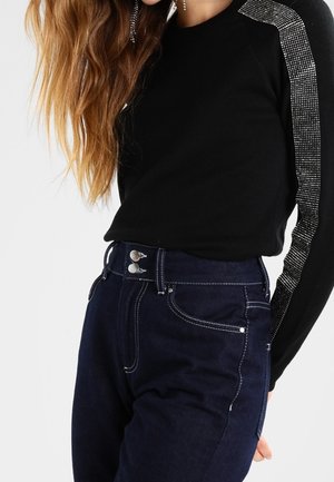 Haut noir à manches longues avec des accents en cristaux sur l'épaule, associé à un jean taille haute bleu foncé avec une fermeture à bouton et des coutures contrastées.