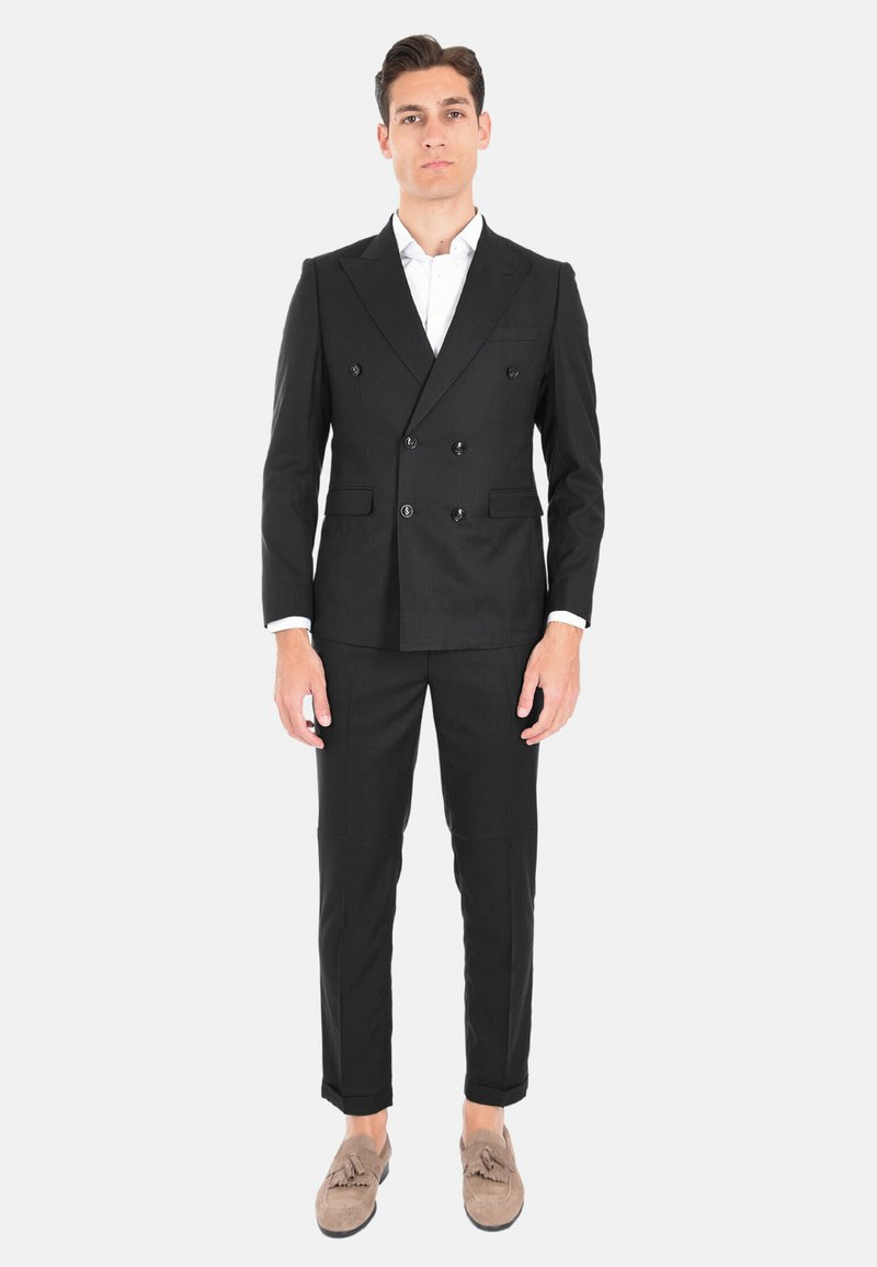 Abito nero doppiopetto con vestibilità su misura, caratterizzato da un rever a punta e tasche laterali. Abbinato a mocassini in suede beige.