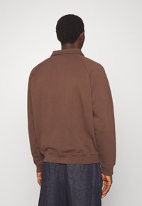 Felpa marrone con colletto e maniche raglan, caratterizzata da un tessuto testurizzato. Indossata con pantaloni in denim scuro.