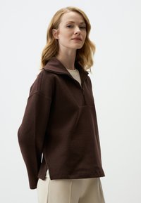 Pullover marrone realizzato in tessuto testurizzato, con colletto a mezza zip e spacchi laterali per facilità. Indossato sopra una camicia di colore chiaro.