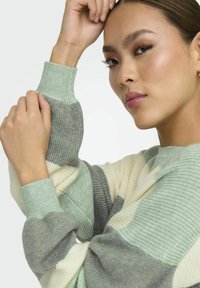Pull multicolore avec des poignets côtelés et des motifs en vert menthe, gris clair et crème. Texture douce avec un design à coupe décontractée.