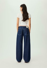 Široke temne jeans hlače z rumenim šivanjem, pet žepov in blagovno oznako na zadnjem pasu. Uporabljene z oprijetim belim zgornjim delom.