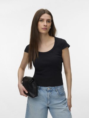 Young woman with long brown hair wearing black t-shirt and light blue jeans, holding a black clutch bag against a plain white background.