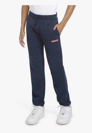 Person iført marineblå Levi's sweatpants med rødt logo, hvide sneakers og en hvid skjorte, højre hånd i lommen, står oprejst.