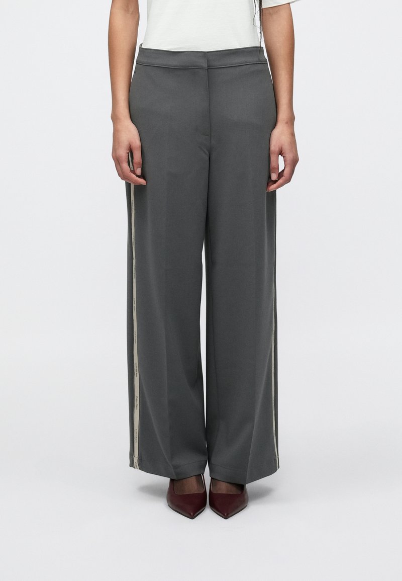 Γκρι παντελόνι με φαρδιά γραμμή κατασκευασμένο από λείο ύφασμα, με μπεζ λωρίδες στο πλάι με λεπτομέρειες κειμένου, συνδυασμένο με μπορντό γόβες με μυτερή μύτη.