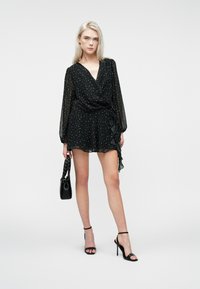 Kvinde i sort polkaprikket omslagskjole med transparente lange ærmer, holder en sort håndtaske, iført sorte højhælede sandaler, står på hvid baggrund.