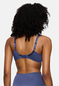 Ženska s kodrasto pričesko, oblečena v modri nastavljivi modrček in ujemajoče se spodnje perilo, prikazana zadaj na bež ozadju.